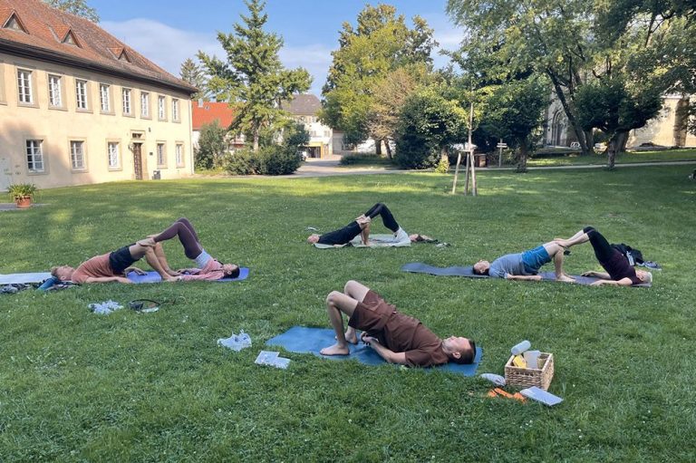 Eine Gruppe von Personen macht gemeinsam Yoga-Übungen auf einer Wiese. Im Hintergrund Bäume und Gebäude.