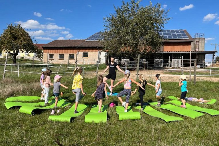 Mehrere Kinder machen gemeinsam Yoga-Übungen draußen auf einer Wiese bei klarem Himmel.